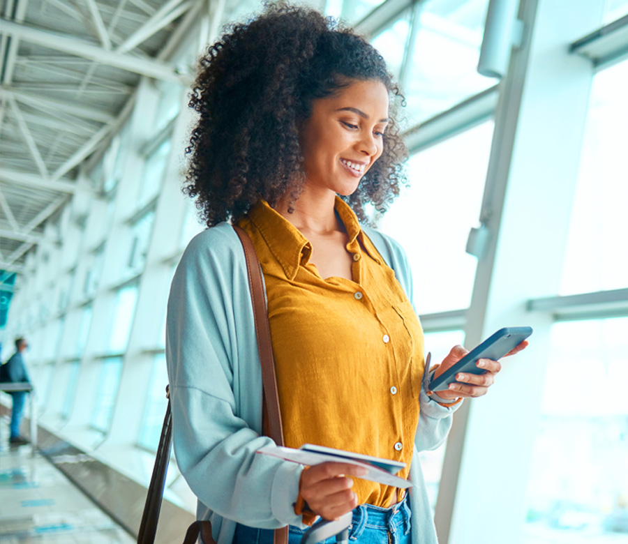 A woman stands at an airport with her boarding pass and smiles at her phone