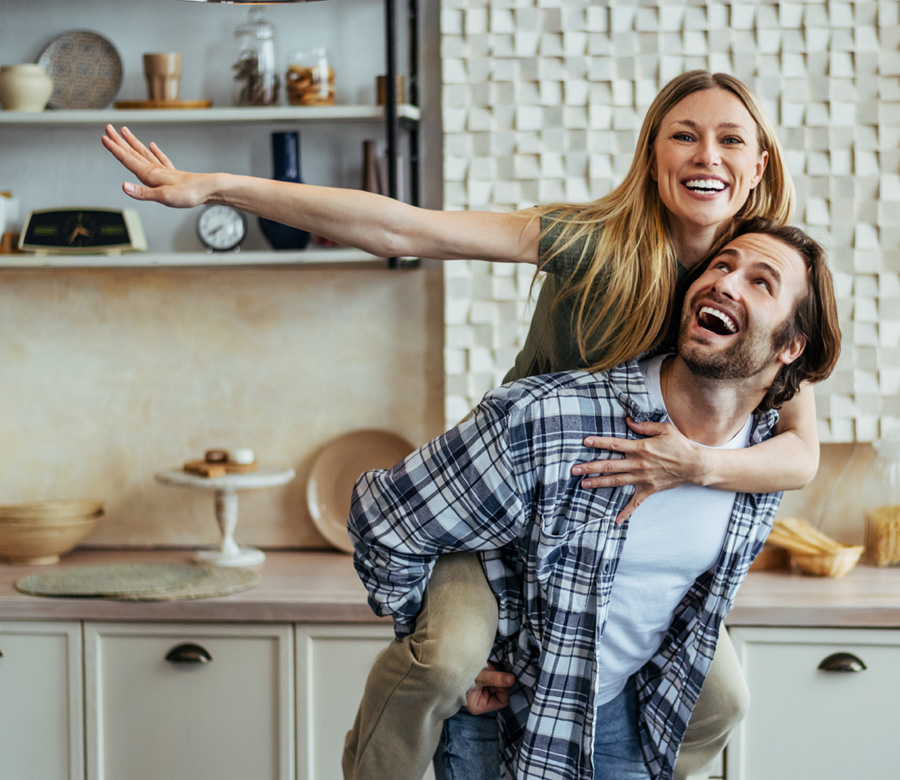 Smiling woman rides piggyback on her husband in their kitchen.