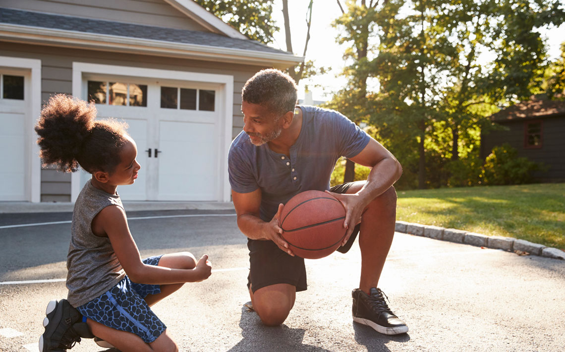 A man and his daughter kneel together in their driveway, the dad is holding a basketball.