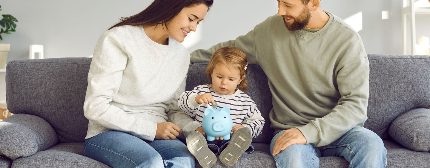 Image of a mom, dad, and child sitting on a couch putting money into a piggy bank.
